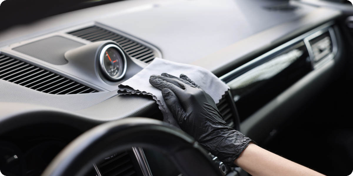 Eine Person trägt schwarze Handschuhe und reinigt das Armaturenbrett eines Autos mit einem grauen Mikrofasertuch.