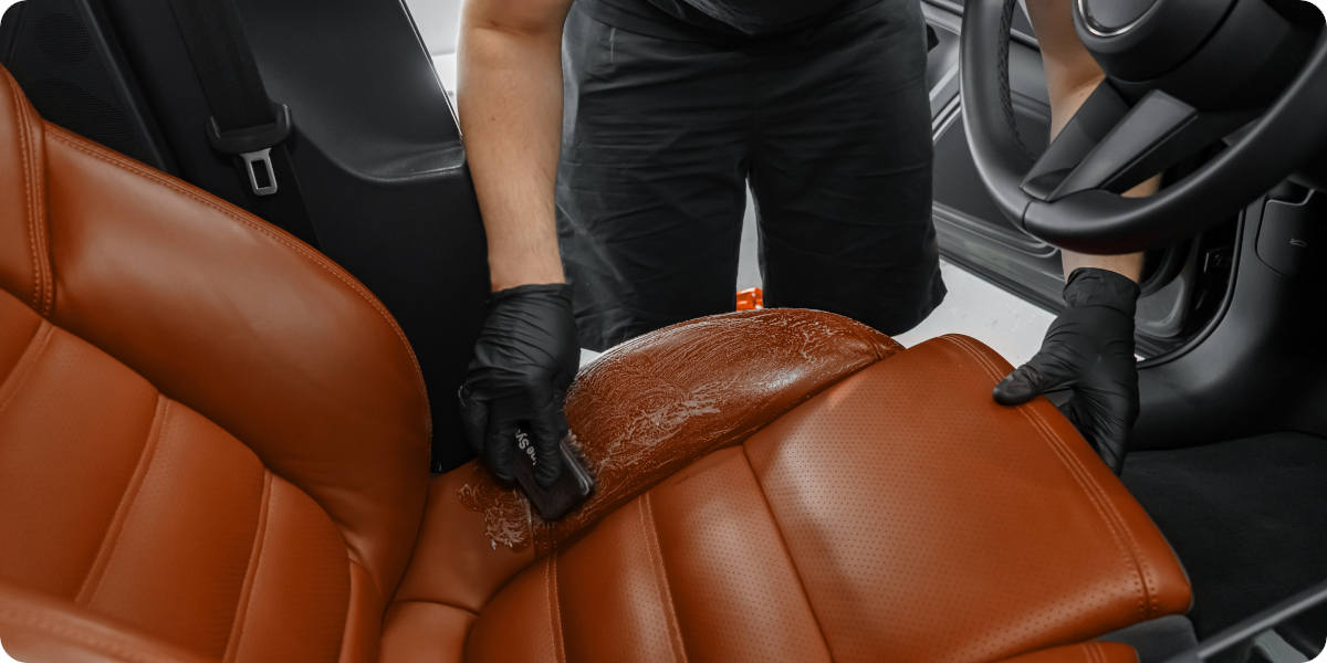 Brauner Ledersitz im Auto wird mit Pflegemittel und Schwamm sorgf&auml;ltig gereinigt.