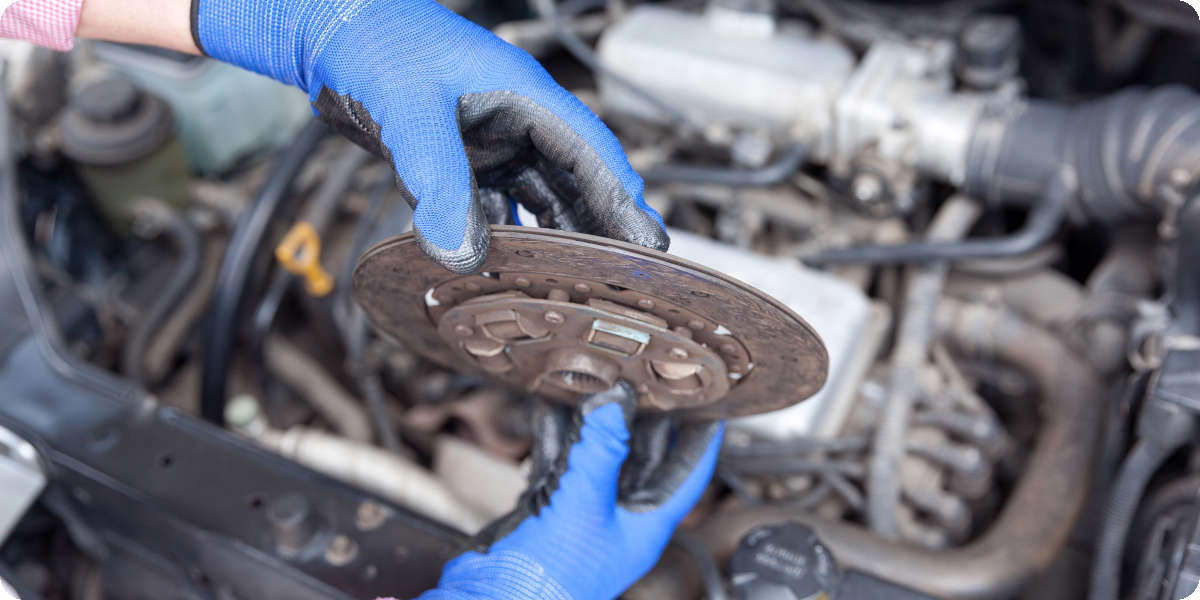 Zwei H&auml;nde mit blauen Gummihandschuhen halten eine verschlissene Kupplungsscheibe &uuml;ber dem Motorblock. 
